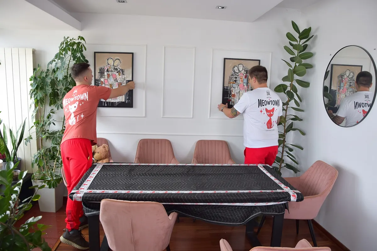 Two Red Cat apartment movers hanging paintings above a blanket-wrapped dining table