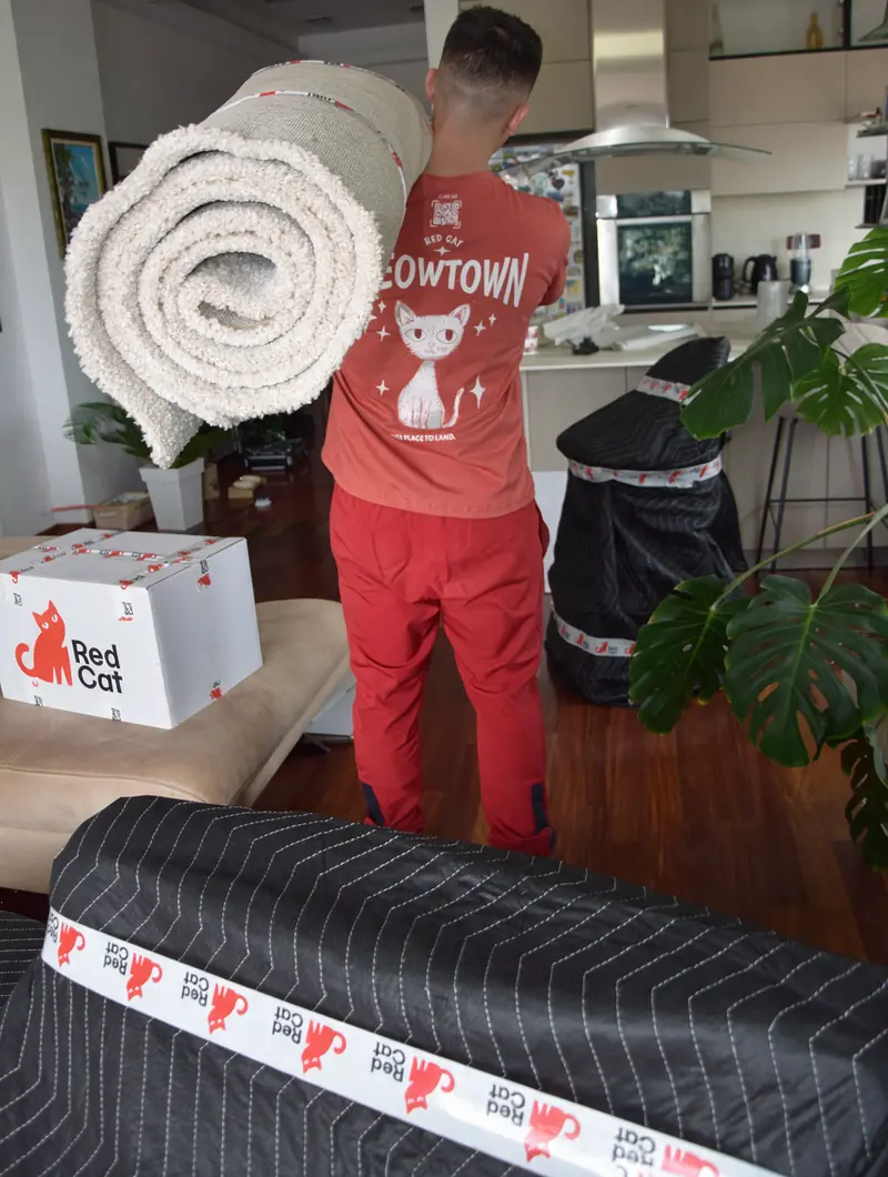 A Red Cat mover carrying a rolled rug past wrapped furniture
