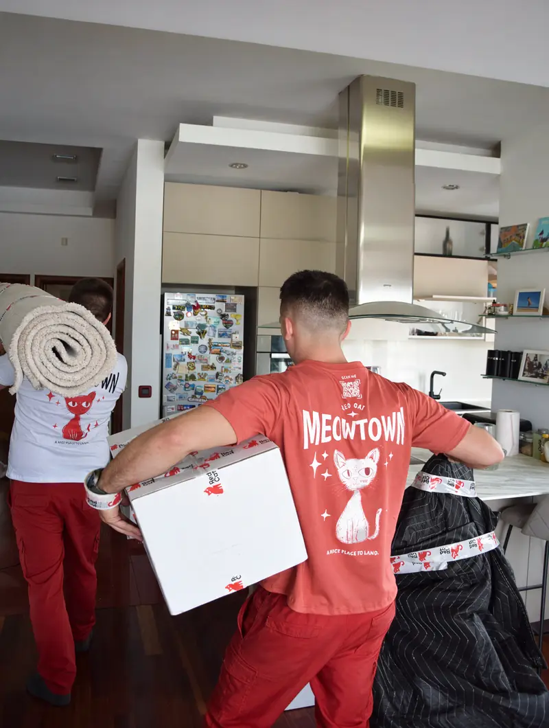 Red Cat movers carrying a box and a rolled rug through a kitchen