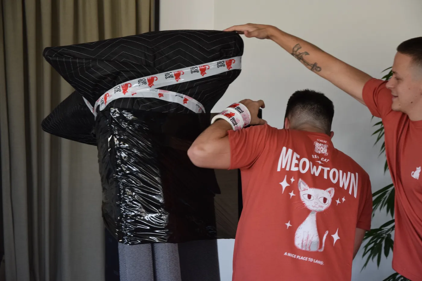 Two Red Cat movers wrapping a tall piece in moving blankets and Red Cat tape