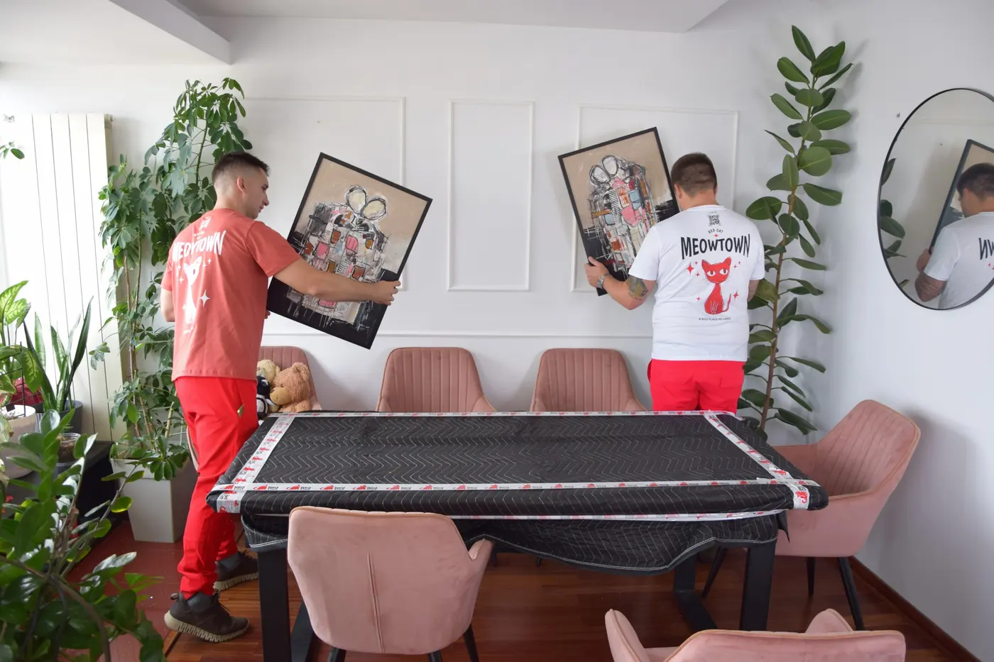 Two Red Cat movers taking paintings down over a blanket-covered dining table
