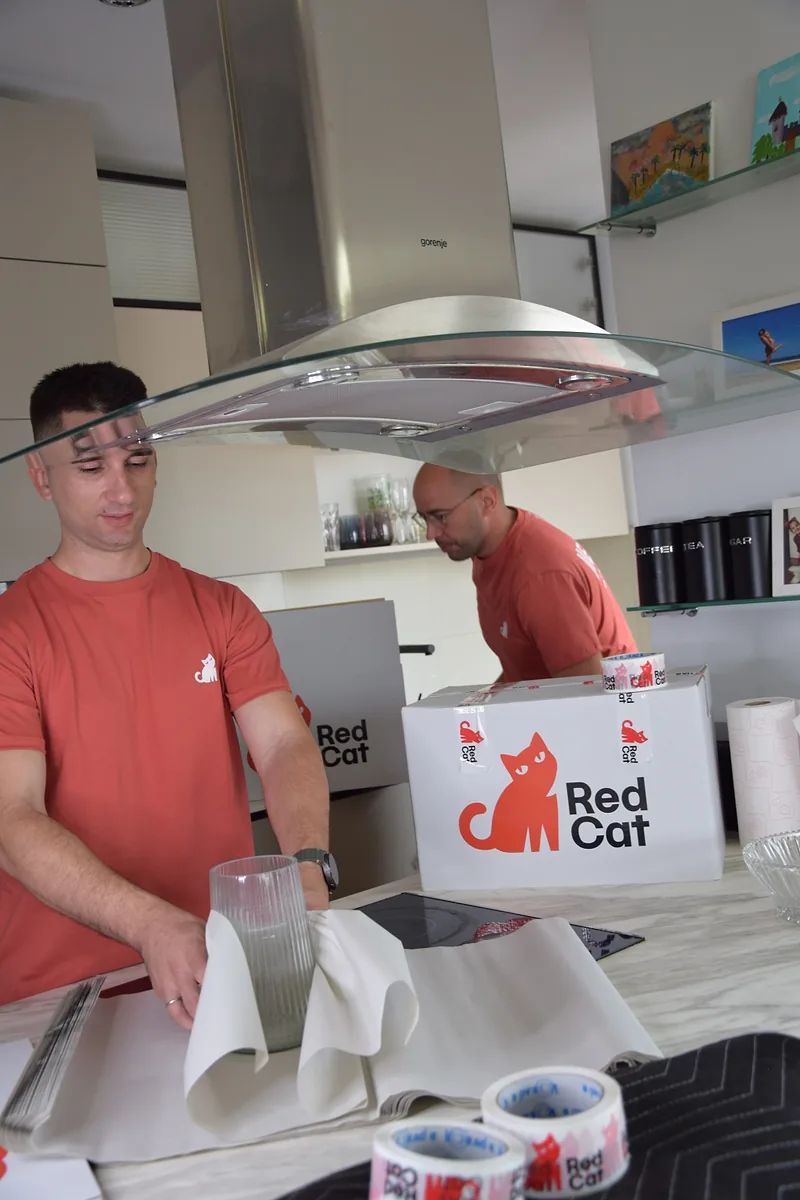 Two Red Cat movers and packers wrapping glassware in a kitchen