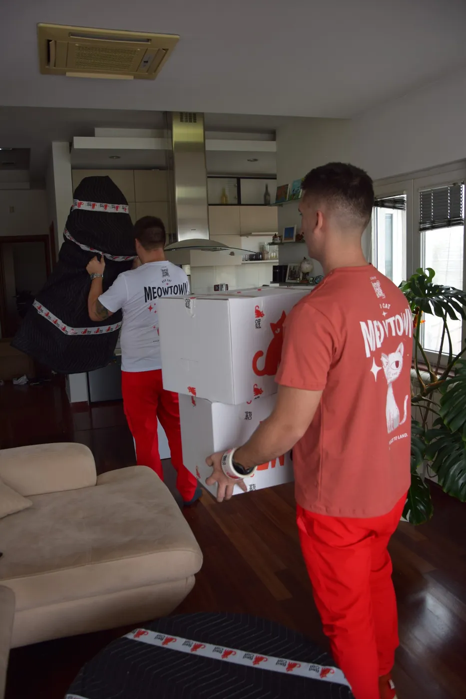 Red Cat movers carrying Red Cat boxes and a blanket-wrapped piece through a living room